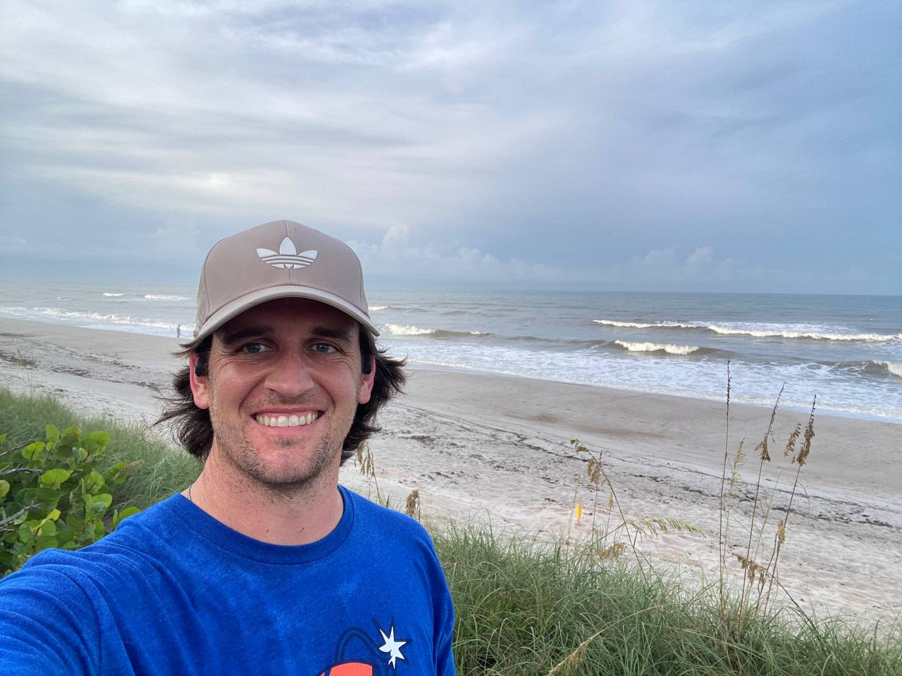 Drew smiling in a ballcap and blue shirt, stopped mid-run to look at the waves on the beach